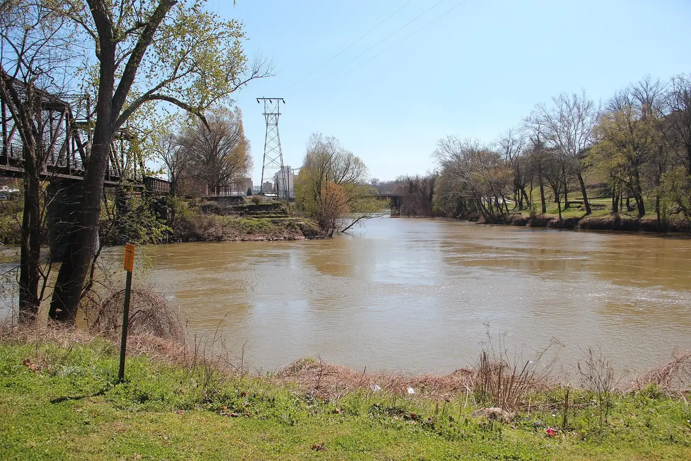 Two brown rivers meeting under a blue sky with bare trees and a transmission tower on the far bank